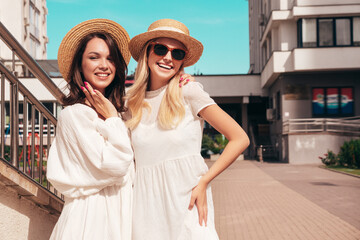 Naklejka premium Two young beautiful smiling hipster female in trendy summer white dress clothes and straw hats. Sexy carefree women walking in street. Positive models having fun, hugging and laughing