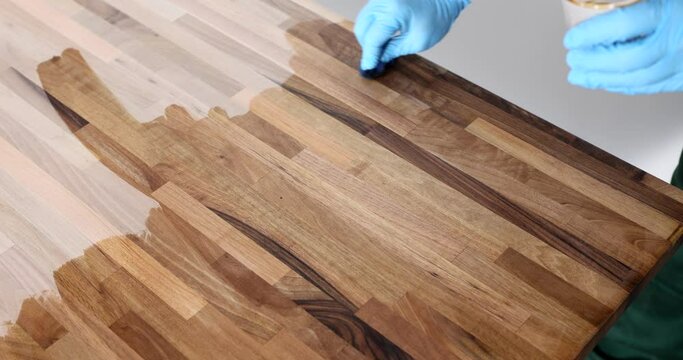 Hands of carpenter in gloves covering wooden surface with protective liquid varnish
