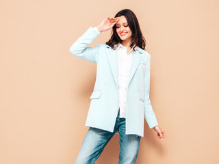 Portrait of young beautiful brunette woman wearing nice trendy blue suit jacket and jeans. Sexy fashion model posing in studio. Fashionable female isolated on beige. Cheerful and happy
