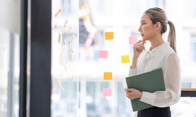 Young Asian woman Creative team use post it notes to share idea sticky note on glass wall. Asian business people design planning and Brainstorming thinking sticky History notes concept.	