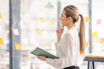 Young Asian woman Creative team use post it notes to share idea sticky note on glass wall. Asian business people design planning and Brainstorming thinking sticky History notes concept.	