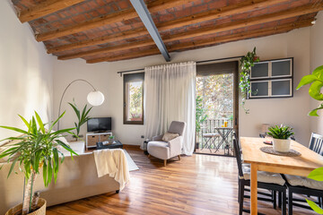 Cozy living area with wood-beamed ceiling with dining table sofas and TV with potted palm tree overlooking the open balcony door. Mediterranean house interior concept