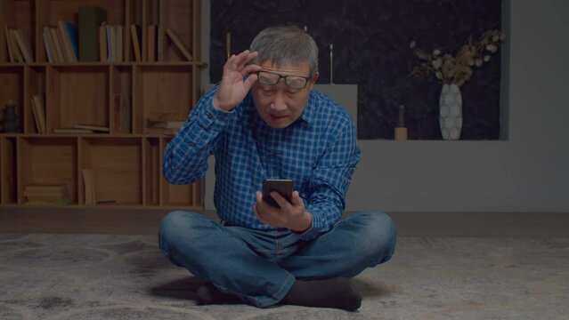 Attractive senior Asian man in eyeglasses having problems with eyesight, squinting eyes and reading message in online chat on cellphone while sitting on floor in domestic room.