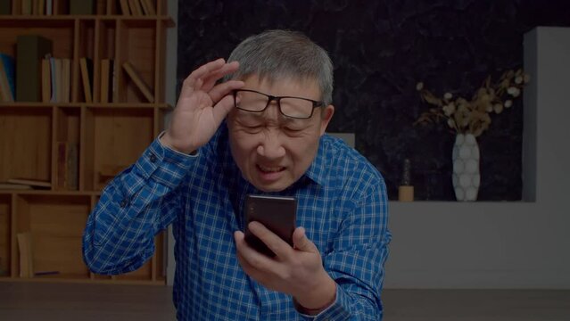 Portrait of handsome retired Asian man in eyeglasses with poor eyesight networking and online chatting on cellphone, squinting eyes and reading message in domestic room.
