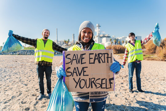 On foreground one happy environmental volunteer smiling and holding a poster cardboard with positive message, save the earth save yourself, at background her friends cleanup trash of the beatch sand - Powered by Adobe
