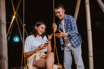 the guy in plaid shirt showing his phone to the woman in white t shirt that sitting on the cradle and pointing on the phone