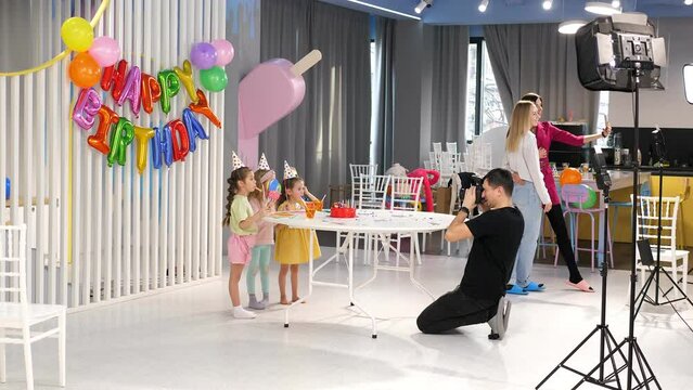 A male photographer is photographing a group of children at a birthday party, children with balloons and caps are standing near a table with a cake.