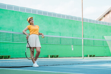 woman in tennis sportswear holding racket and ball leaning on net in tennis court