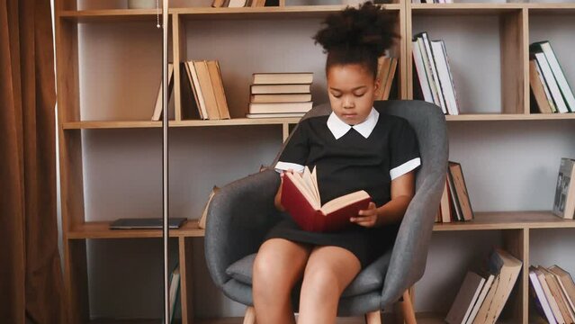 Sitting in the chair and reading the book. Cute african american girl in school uniform is at home library.