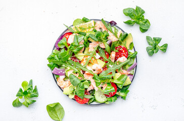 Delicious shrimp salad with avocado, cherry tomatoes, arugula, lamb lettuce, cucumber, red onion and sesame seeds on white table background. Top view