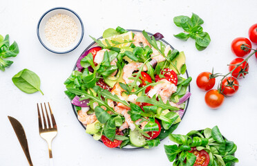 Delicious shrimp salad with avocado, cherry tomatoes, arugula, lamb lettuce, cucumber, red onion and sesame seeds on white table background. Top view