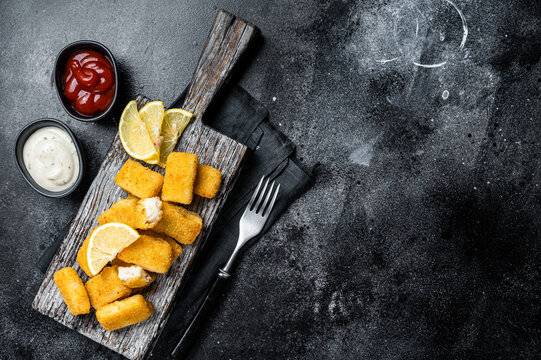 Golden roast Fish Sticks Fingers with sauce on a wooden board. Black background. Top view. Copy space
