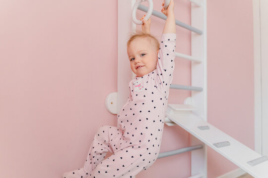 A Caucasian girl in soft pink pajamas does sport at gymnastic rings