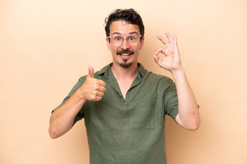 Young caucasian man isolated on beige background showing ok sign and thumb up gesture