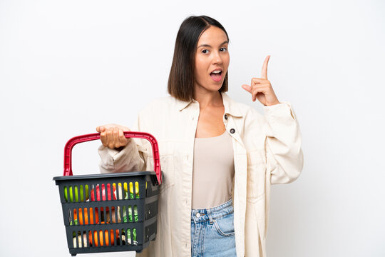 Young woman holding a shopping basket full of food isolated on white background thinking an idea pointing the finger up