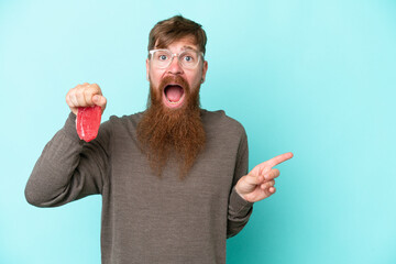 Redhead man with long beard holding a piece of meat isolated on blue background surprised and pointing finger to the side