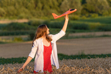 Happy young woman with toy airplane in hands in middle of wheat field, in red dress and white shirt Concept of cheap air travel