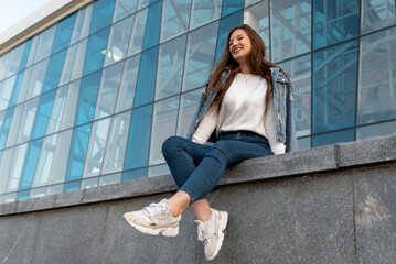 Laughing stylish girl resting on summer city street. Beautiful student girl sitting near modern building