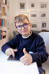 Bambino biondo con gli occhiali , vestito in blu e seduto davanti a una scrivania, utilizza un...