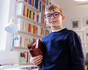Bambino biondo con gli occhiali vestito sta in piedi con dei libri in mano dentro lo studio