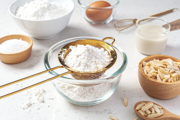 Sifting flour with gold sieve in glass bowl with baking ingredients on marble kitchen table