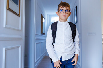 Bambino biondo con gli occhiali e lo zaino in spalla sta  in piedi nel corridoio di casa