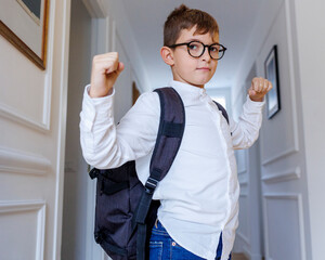Bambino biondo con gli occhiali e lo zaino in spalla in piedi nel corridoio di casa mostra i...