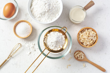 Sifting flour with gold sieve in glass bowl with baking ingredients on marble kitchen table