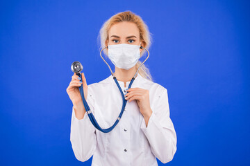 Portrait of a young female doctor in a protective mask on a blue background.A stethoscope in the hands of a doctor