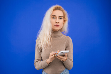A young woman with curly hair dials a number on a smartphone on a blue background in the studio