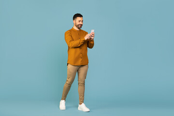 Full length shot of italian man walking and using cellphone chatting with friends blue background