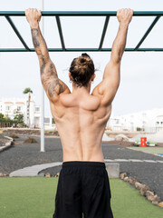 Muscular shirtless caucasian man from behind doing pull ups at the calisthenics park