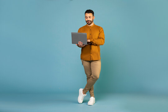 Full body length shot of young european man in casual outfit using laptop computer blue background, free space