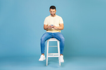 Young man using smartphone, chatting online, browsing social media, sitting on chair on blue...