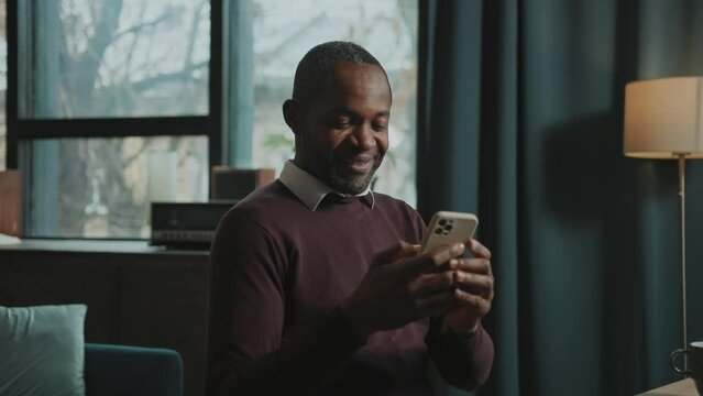 Horizontal footage of African American smiling businessman in brown sweater looking at screen of smartphone, texting in messenger in cozy atmosphere of home. Modern furniture on background