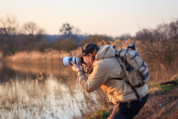 Photographer with camera, telephoto lens and backpack taking picture at lake during sunset. Hobby and outdoor leisure activity - obrazy, fototapety, plakaty