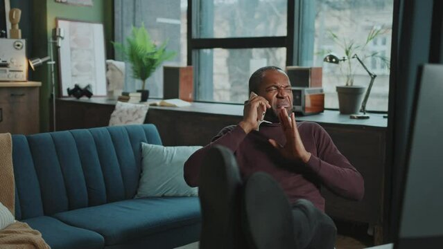Footage of confident busy African American businessman sitting at workplace with legs on desk in cabinet at home. Relaxed carefree man holding smartphone in hand and talking to business partner