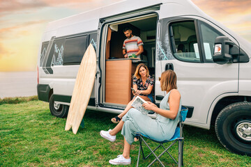 Group of friends traveling in a camper van. Two women talking in front of camper van while young man cooks © David Pereiras