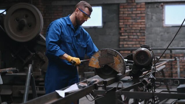 Factory worker that is in blue uniform and in protective glasses is cutting metal.