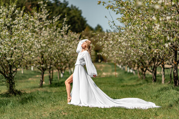 Obraz na płótnie Canvas Blond blooming garden. A woman in a white dress walks through a blossoming cherry orchard. Long dress flies to the sides,