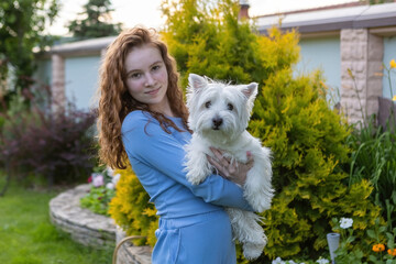 A young red-haired girl holds a white dog in the outdoors in the summer