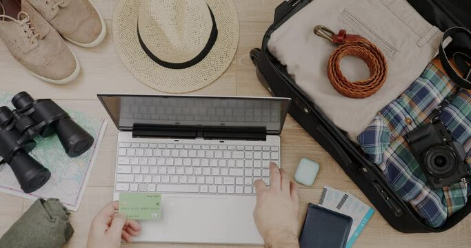 Top view of hand with credit card using laptop buying holiday trip with packed suitcase and travel objects visible against wooden background