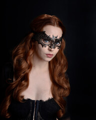 Masked close up portrait of beautiful woman with long red hair wearing sheer corset top, and black bat wing mask.  Isolated on dark studio background with. Moody silhouette lighting.