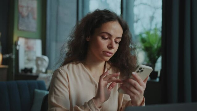 Portrait of pretty thoughtful woman sitting in front of laptop and holding smartphone in hands. Footage of nice girl touching screen, scrolling through social media feed and looking away