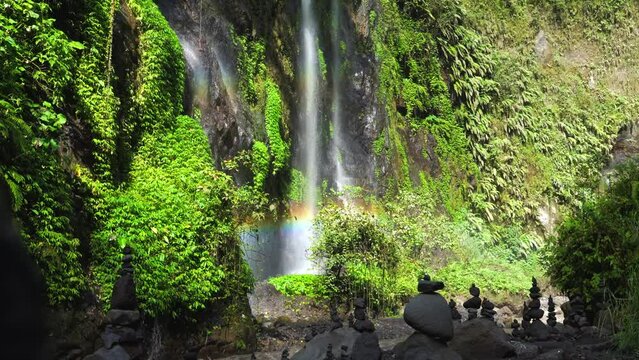 Rock cairns stacked at the base of a tropical waterfall that creates a rainbow in the canyon of a mystical rainforest, East Java, Slow motion panning up