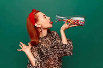Beautiful young redhead woman in stylish clothes eating multicolored candies over dark green studio background. Concept of food pop art photography. Complementary colors. Copy space for ad, text