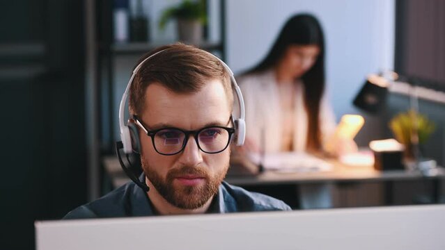 Man in headphones with microphone is working by a computer in the office. Woman is behind.