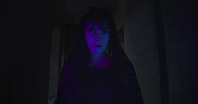 Creepy mysterious goth girl with lace hood and purple lighting on her face staring directly into camera, slow motion