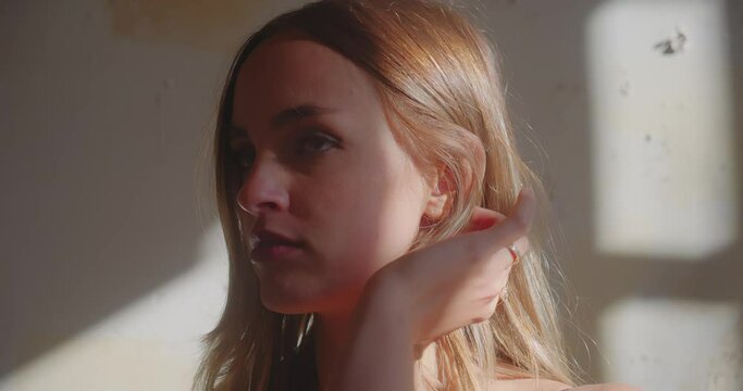 Young blonde caucasian woman putting hair behind ear with hand in her room, sunlight shadows caused by window in the background