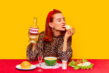 Food pop art photography. Beautiful redhead woman sitting at table against yellow background and having dinner, drinking vodka with onion and salad. Complementary colors. Copy space for ad, text